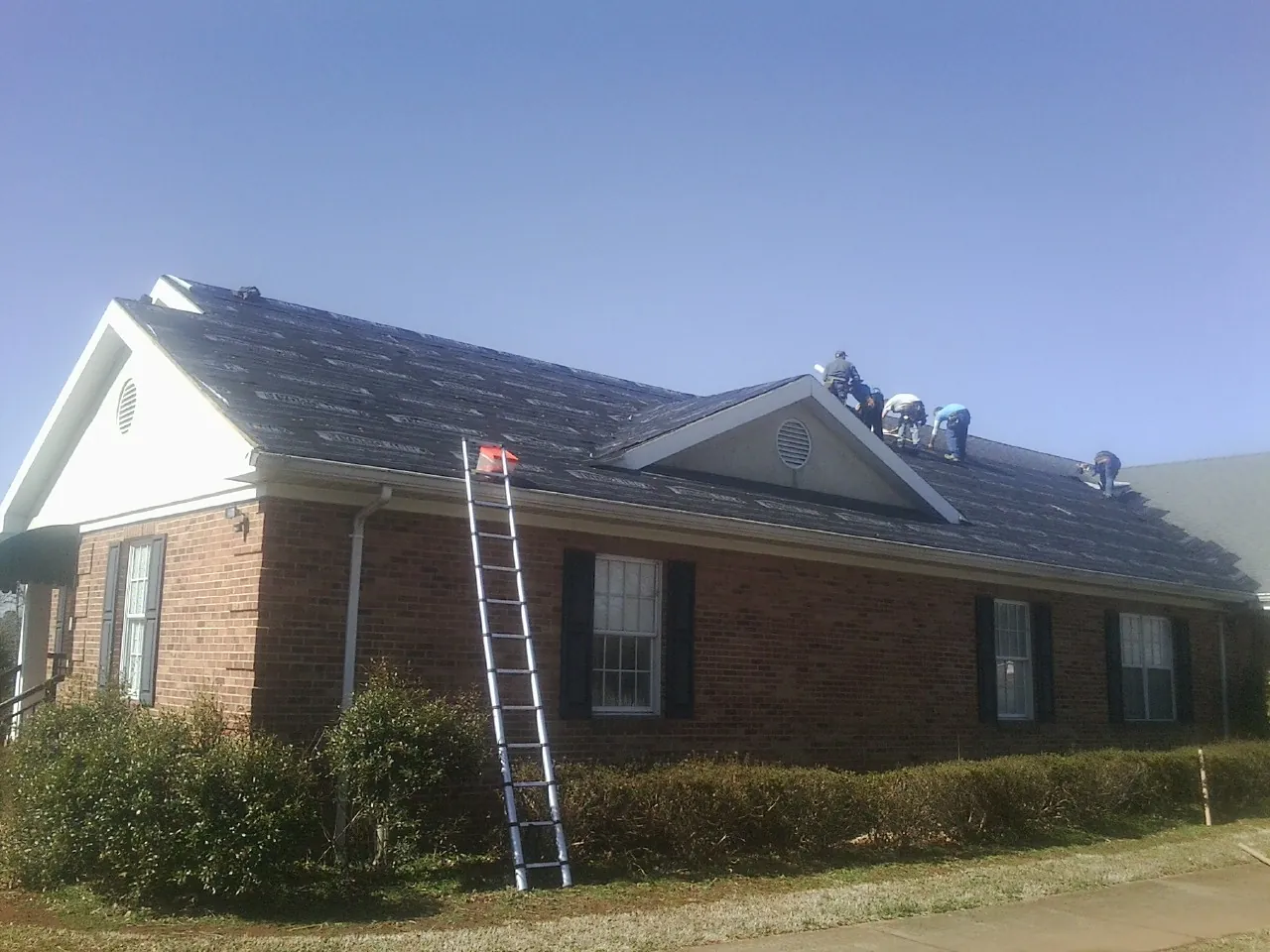 Crew tearing shingles off a brick ranch roof with a ladder set — Metal Roof Repair work by our crew in New Hampton