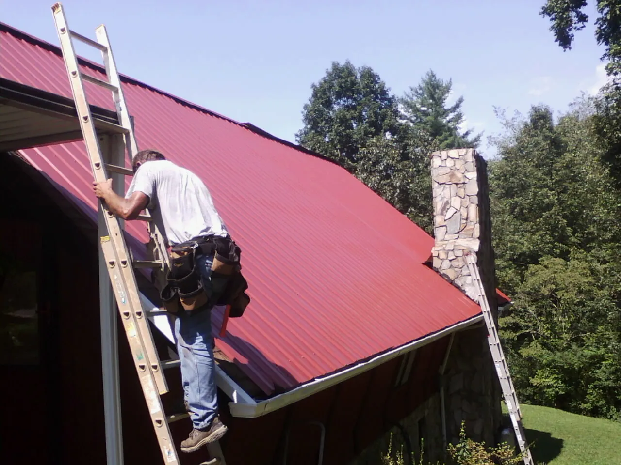 Roofer on a ladder beside a red metal roof and stone chimney — Soffit & Fascia Repair by Roofing Brothers in New Hampton